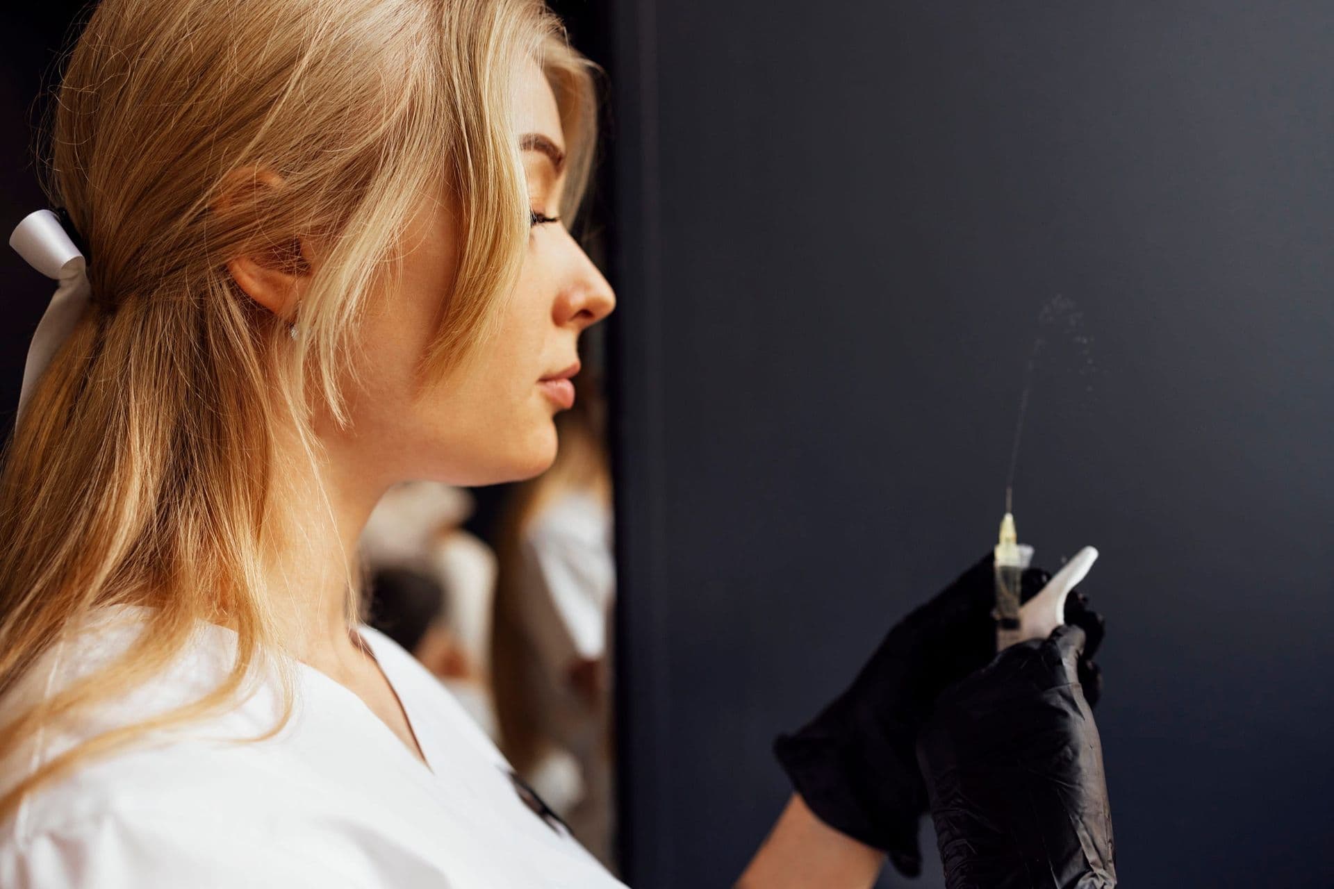 A clinician in black gloves holds a syringe against a dark wall